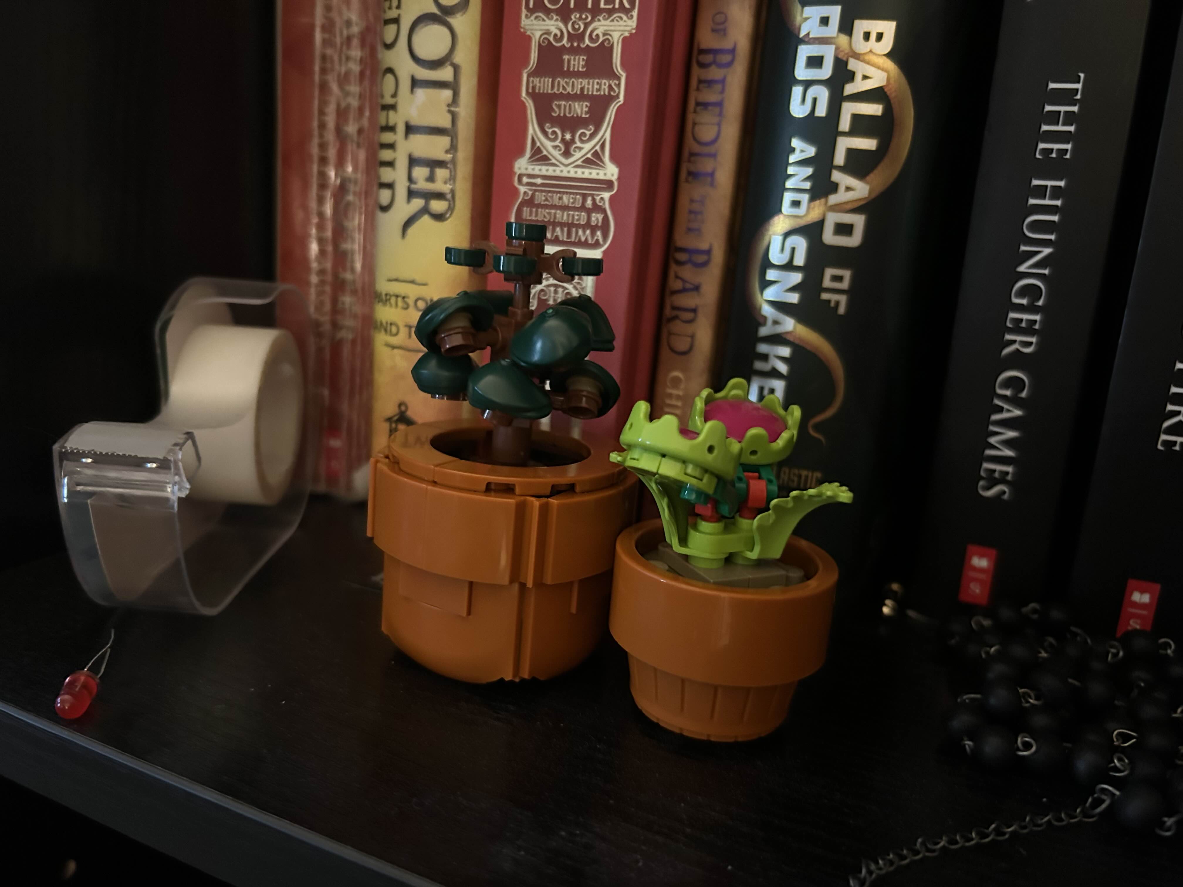 Image of two LEGO flowers sitting on a bookshelf. They are in matching brown pots. The larger, medium sized on is a tall bush with large round leaves. The smaller plant is a venus fly trap.